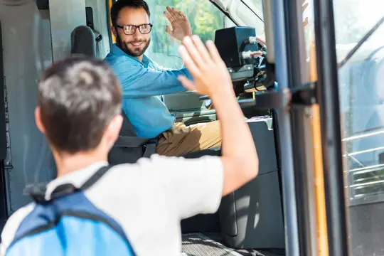 Bilde av en skolegutt som er på vei inn i skolebussen og hilser på buss sjåføren.