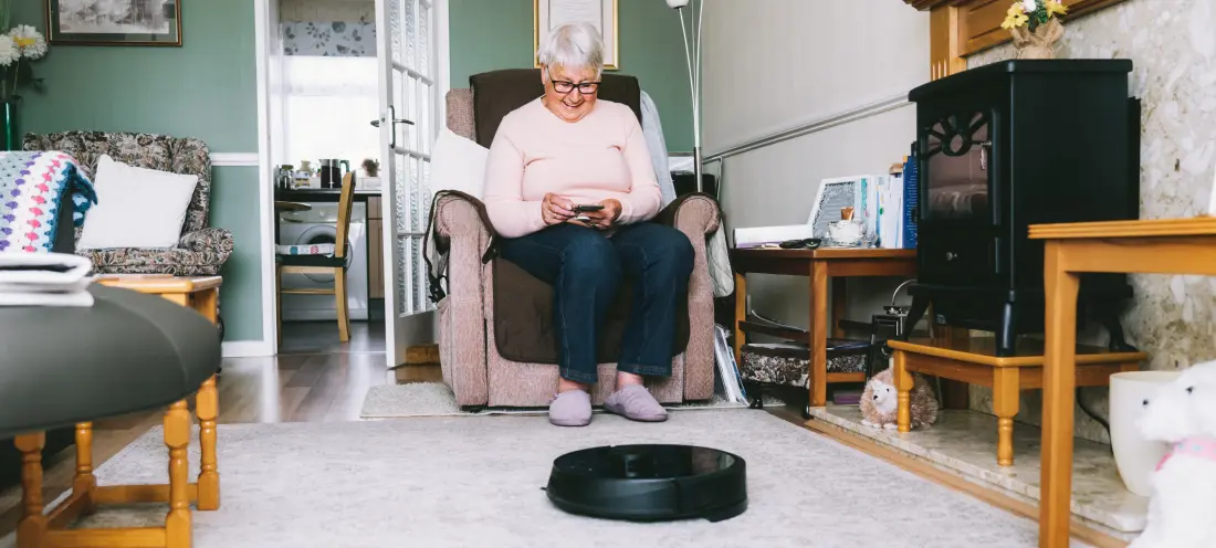 Eldre dame som sitter hjemme med en mobiltelefon og robotstøvsuger.